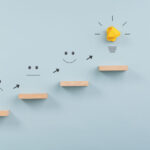 Business vision, idea and development concept - steps made of wooden pegs leading to a lightbulb. Over light blue background.